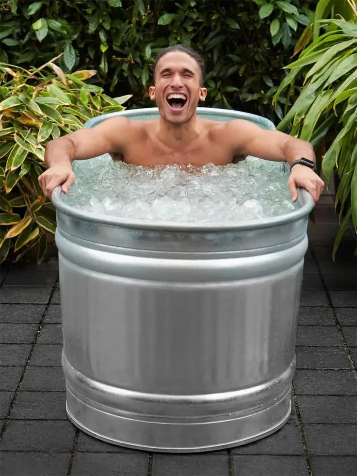 Man smiling while sitting in an ice-filled metal tub during a cold plunge in a garden setting